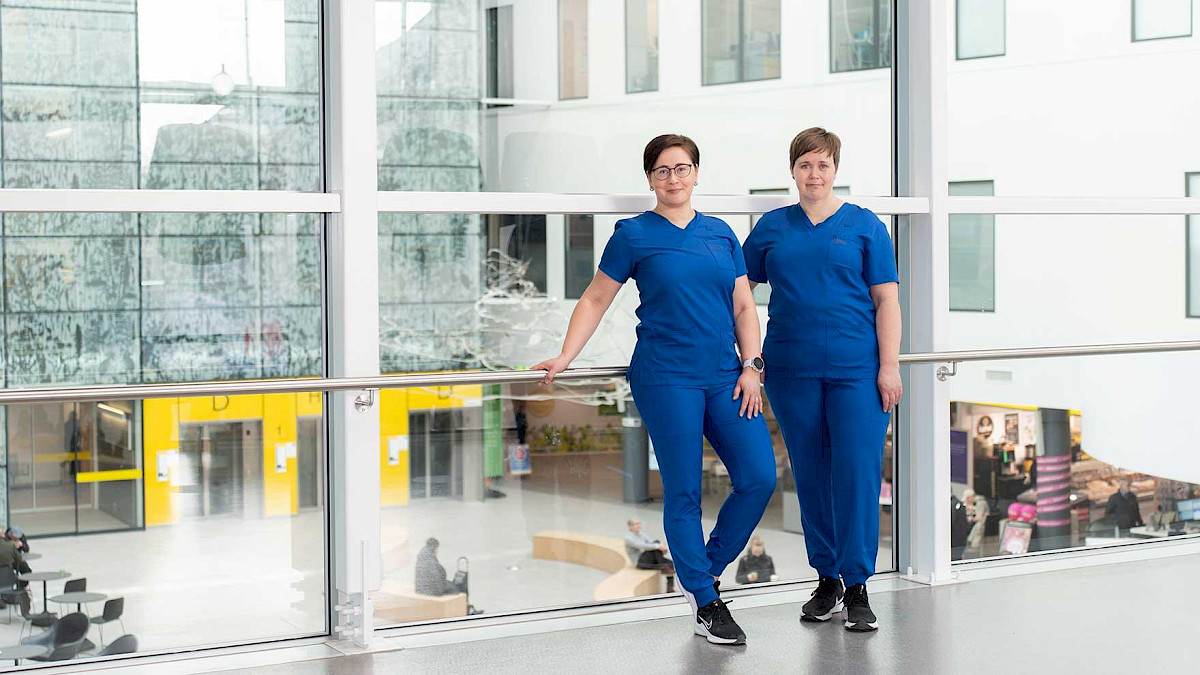 Kaksi siniasuista tutkimushoitajaa kuvattuna Tampereen yliopistollisen sairaalan käytävällä, taustalla lasiseinän takana avautuvat avarat aulatilat.